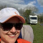 A lady wearing sunglasses is sitting in a camping chair on a campsite with an A Class Motorhome in the background