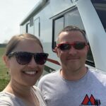 A smiling couple stand in a field, next to an A Class Motorhome