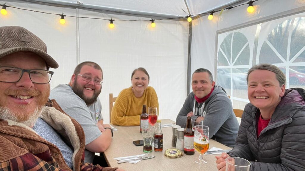 5 people are ast around a table in a pub garden, smiling. There are drinks on the table