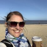 A lady is holding a cup of takeaway coffee and smiling, standing on the beach with the sea behind her