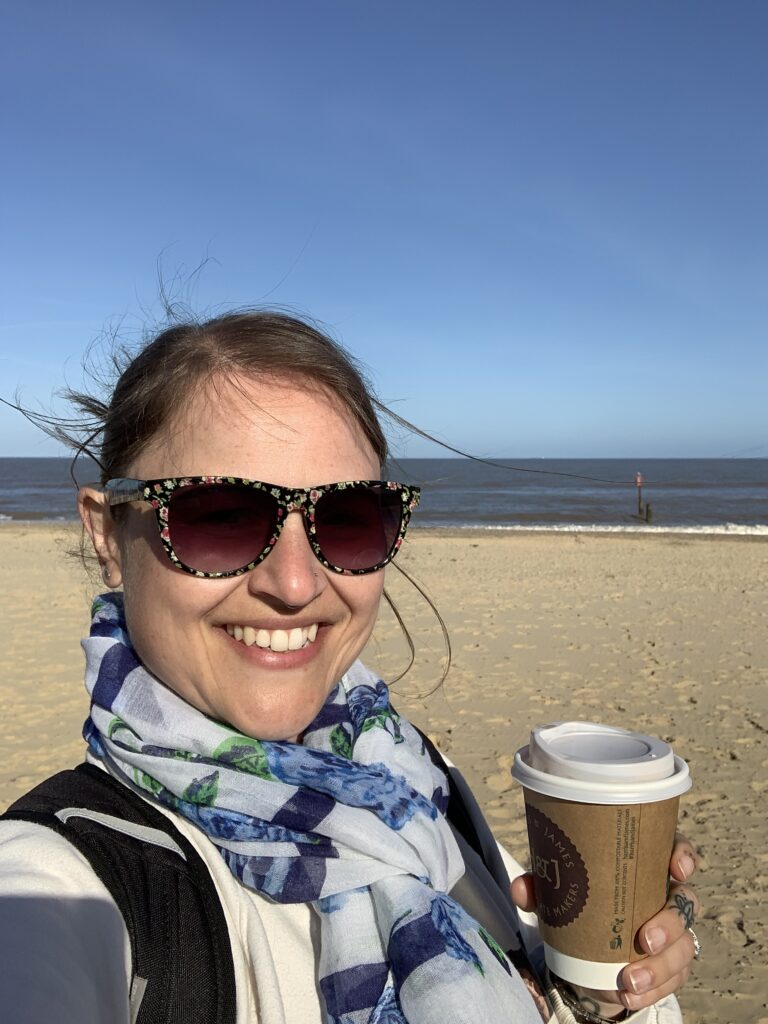 A lady is holding a cup of takeaway coffee and smiling, standing on the beach with the sea behind her
