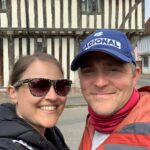 A smiling couple stand in front of a Tudor Style building with cream masonry and brown timber frame