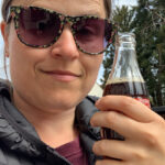 A lady wearing sunglasses is holding a glass bottle of Coca Cola