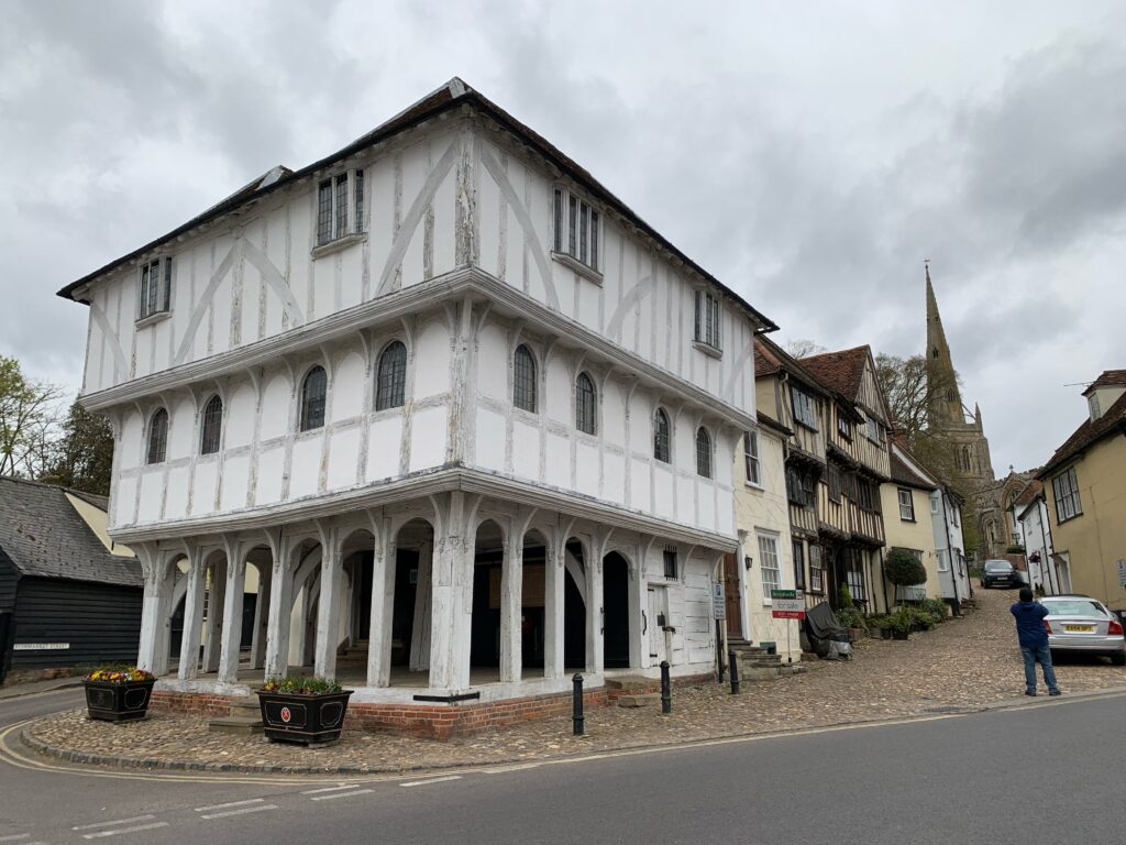 The Thaxted Guildhall. It is 3 stories high, with faded timber framework and white masonry in between. An open air market space if on the lower level and the two levels above are jettied