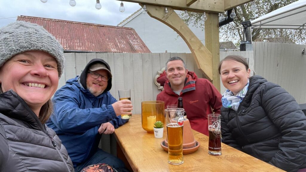 2 men and 2 women are sat around a table in a pub garden. All are smiling and one is raising a glass to cheers