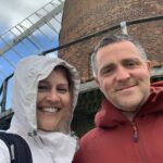 A man and woman stand smiling, underneath a windmill