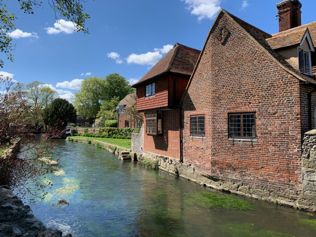 A red brick house sits on the edge of the river stour. The view is looking up the river and there are trees in view