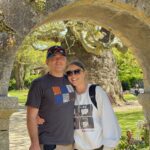 A man and Woman stand smiling, arm in arm, under a stone archway
