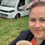 A lady is standing in front of an A class Motorhome holding a cup of coffee and smiling. She has a pretty red headband on