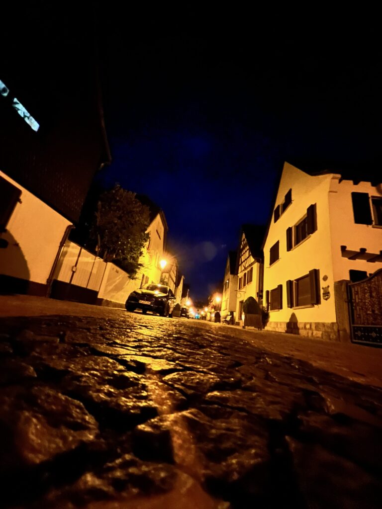 the viewpoint of a German street, the photo is taken from a low angle, from just above the cobbles. The street lamps lights up the rows of houses on either side 