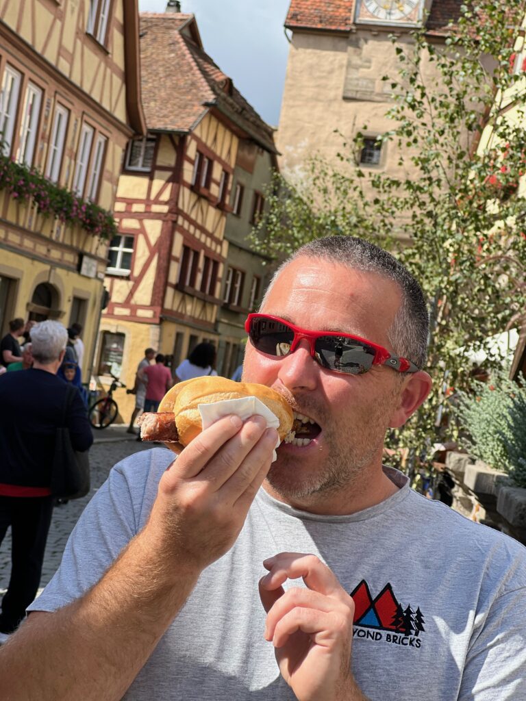 A man eats a bratwurst. The German buildings of Rothenburg Ob Der Tauber are visible in the background