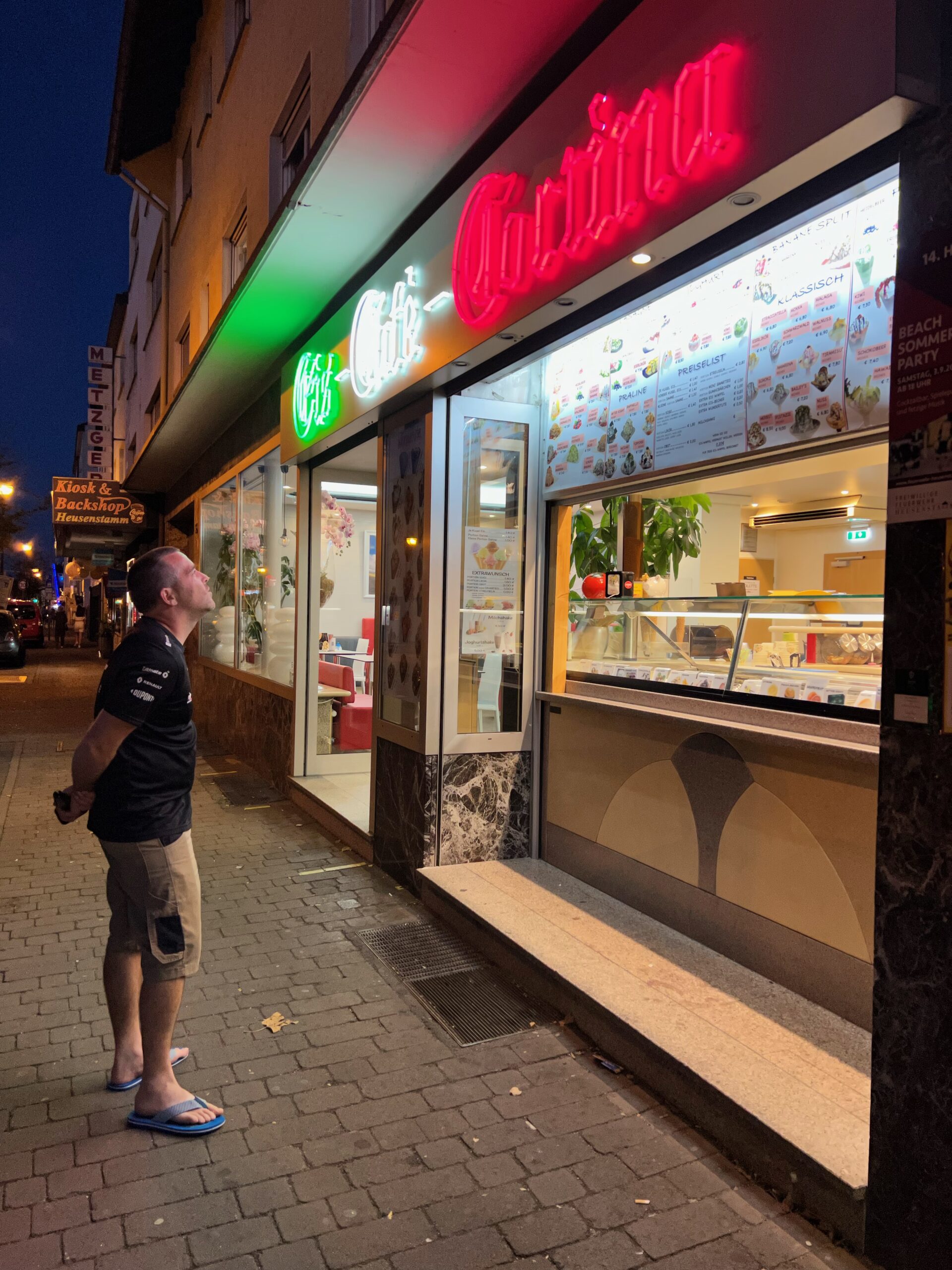A man stands on the pavement of a German street, looking up at the lit menu of an ice cream parlour. The red, green and white sign is illuminated above