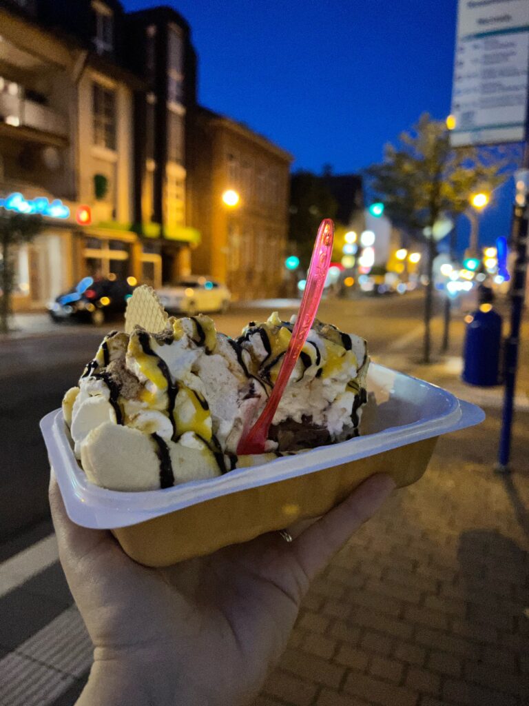 a hand is holding up a banana split ice cream. It has chocolate sauce on top. The street at night is in the background