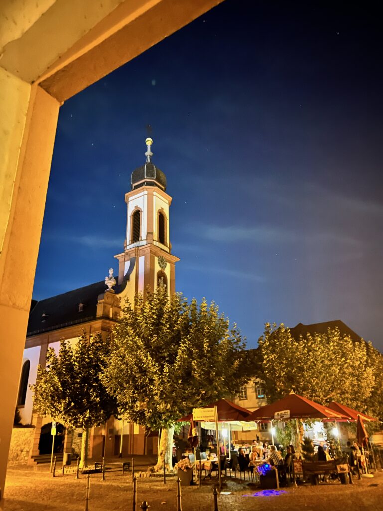 The view is through an archway, just visible in the top let coiner of the picture. through he archway we see the tower of a church against the backdrop of the night sky. There are trees and tables on the ground below of a nearby restaurant 