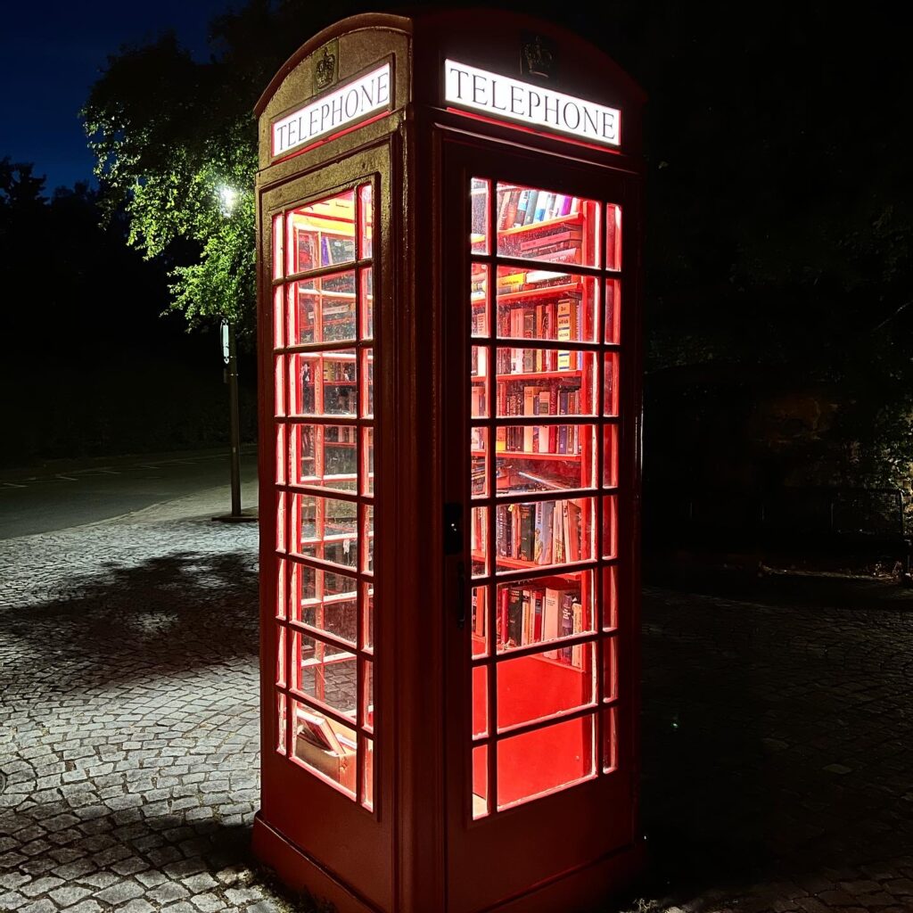 A traditional red British telephone box, it is lit up inside and is full of books
