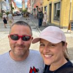 A man and woman stand smiling in the street at Rothenburg ob Der Tauber. ITs a bright day, the man is wearing sunglasses, the lady is wearing a hat