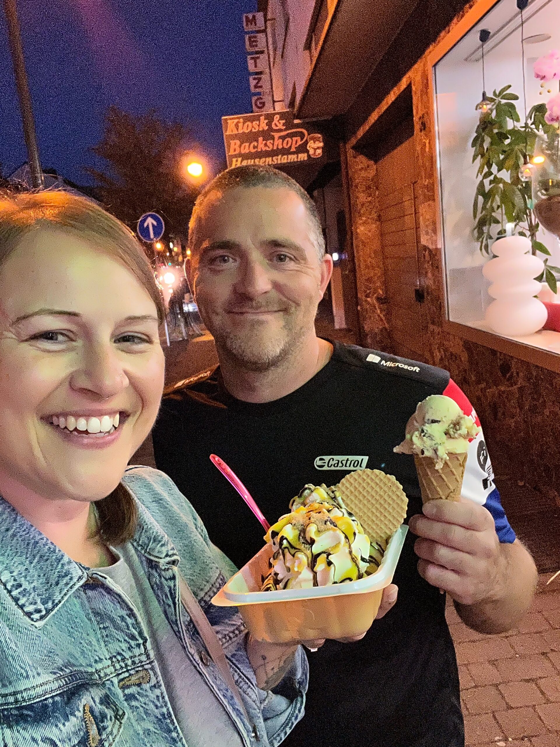 A man and lady are posing together, smiling. the lady is holding a banana split desert in a tray, the man is holding an ice cream cone. They are staying on the pavement at night in a town centre street