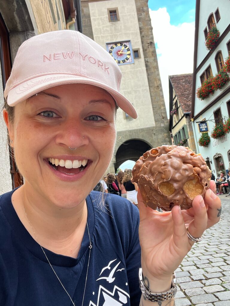 A lady holds up a snowball, a Bavarian shortcrust pastry that is deep friend. It is large and round and covered in chocolate.