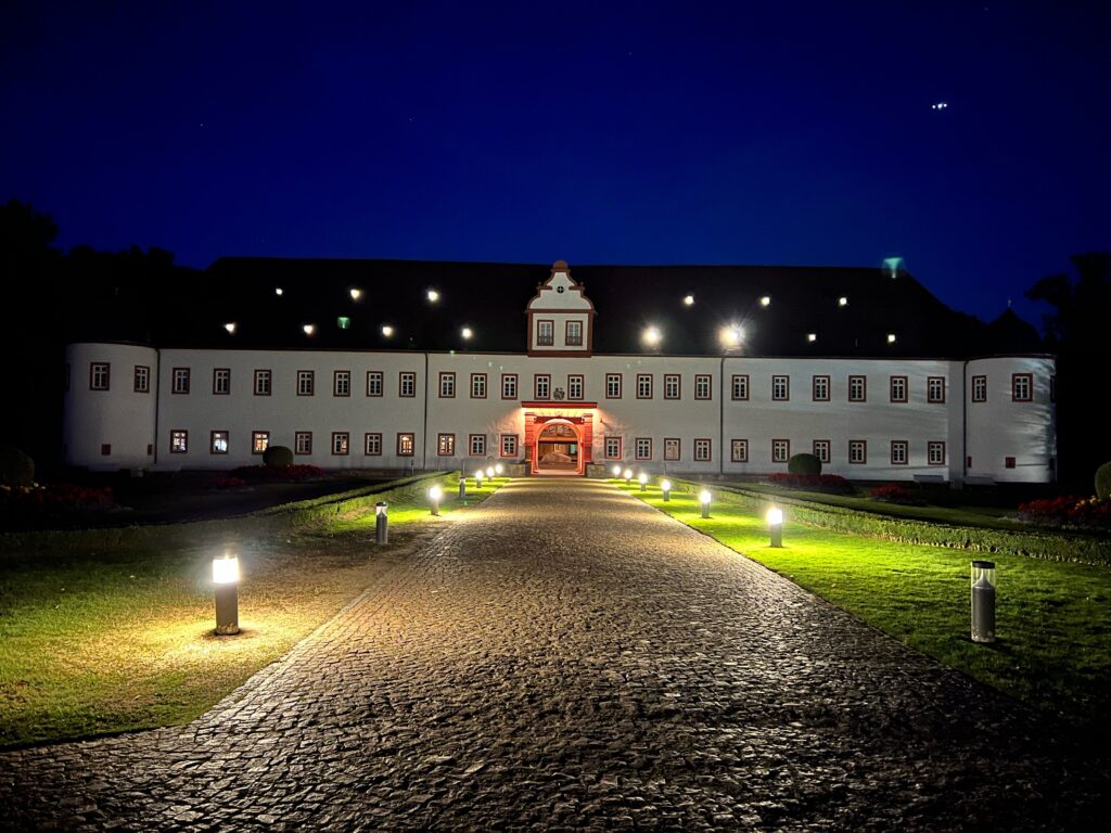 A cobbled path extends up to a white rennaisance castle. It is night time, the path is lit with lights. There is grass either side of the path.