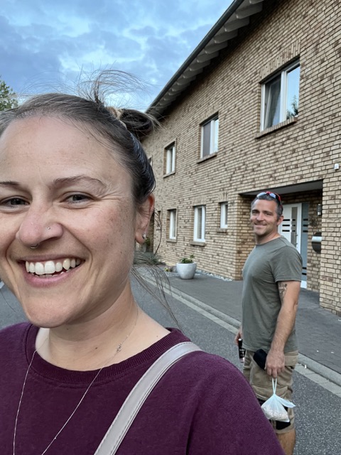 A man and lady are smiling as they pose for a picture down a residential street in Germany, the lady is in front of the man, the man is behind her, visible over her shoulder 