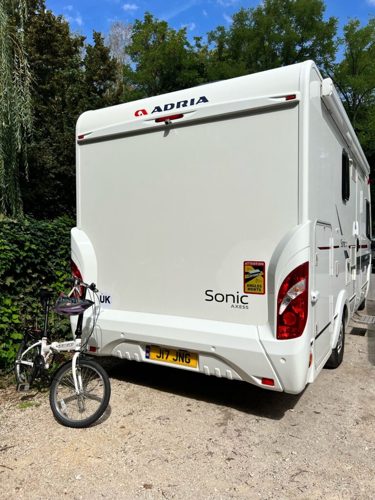 The back of an A class motorhome is in the centre of the shot, parked in a Stellplatz. There is a folding bike stood up behind it