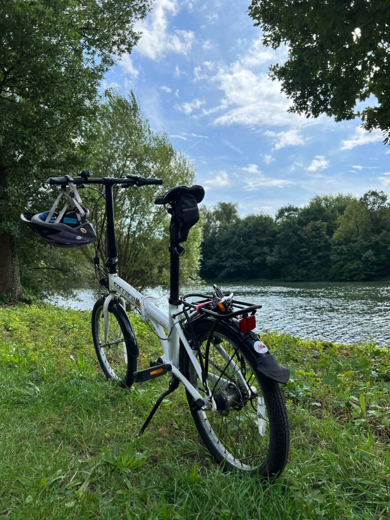 a folding bike is stood on the bank of a river, with a helmet hanging from the handlebar. There are lush green trees framing the picture
