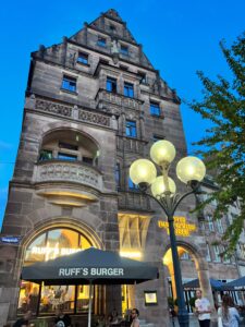 A beautiful building is centre shot, it is tall and narrow with a pointed roof. It is old in character and has a balcony and the bottom is now a burger bar. Picture taken at dusk.