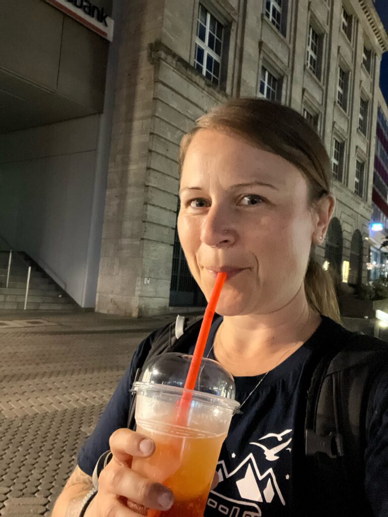 A lady is drinking an aperol spritz from a plastic cup through a straw, she is in nuremberg at night