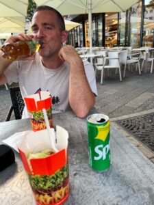 A man sits at a table, he is drinking a bottle of juice and smiling. There is a can of sprite on the table and two doner boxes. The rest of an outside seating area at a cafe is visible in the background