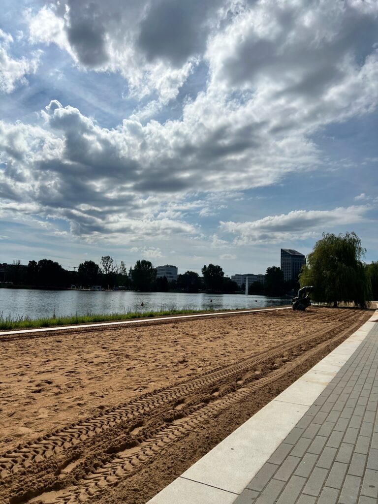 The picture looks out across the river and down the manmade beach along the cycle path in the city if nuremberg. The sand is a dark yellow and the clouds are prominent int he sky, although it is a bight and sunny day
