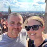 A man and woman are stood on a terrace on a sunny day, the city of nuremberg is visible behind them. The woman is wearing sunglasses and they are both smiling