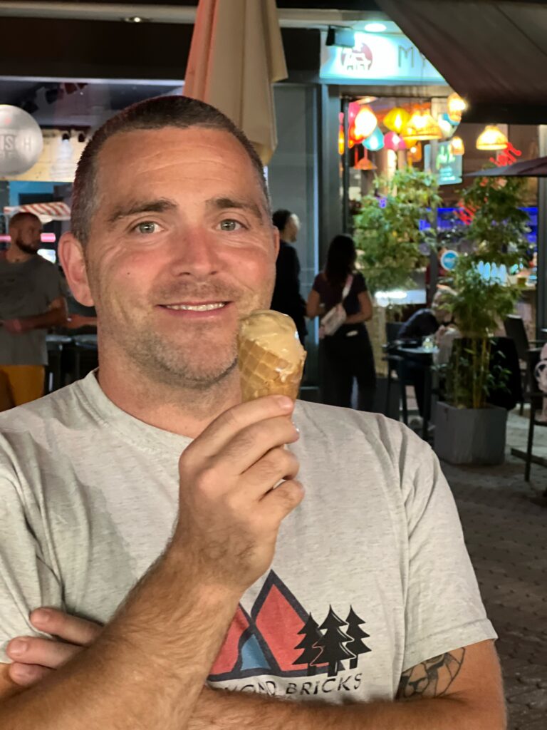 A man is smiling, holding an ice cream. Behind him is the city of Nuremberg at night