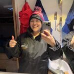 A lady is standing in the bathroom of her motorhome, she is giving a thumbs up and smiling, posing for a selfie. She is wearing a Sahara Challenge heat and coat 