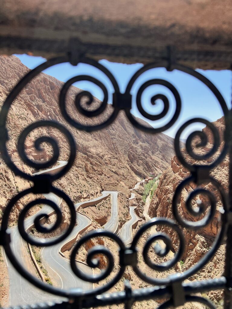 The view is through a metal fence, overlooking the switchback road of the Dades Gorge
