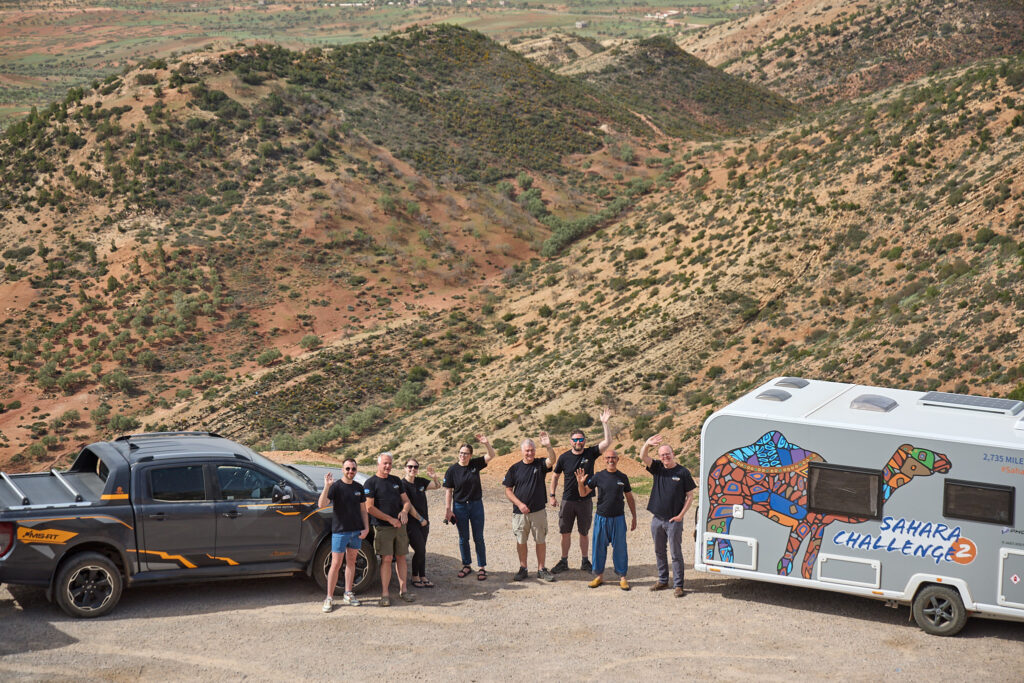 A group of people are stood on the road side, high up above the landscape. There is a ford ranger one side and a caravan the other. The people are stood in between and are waving up at the camera