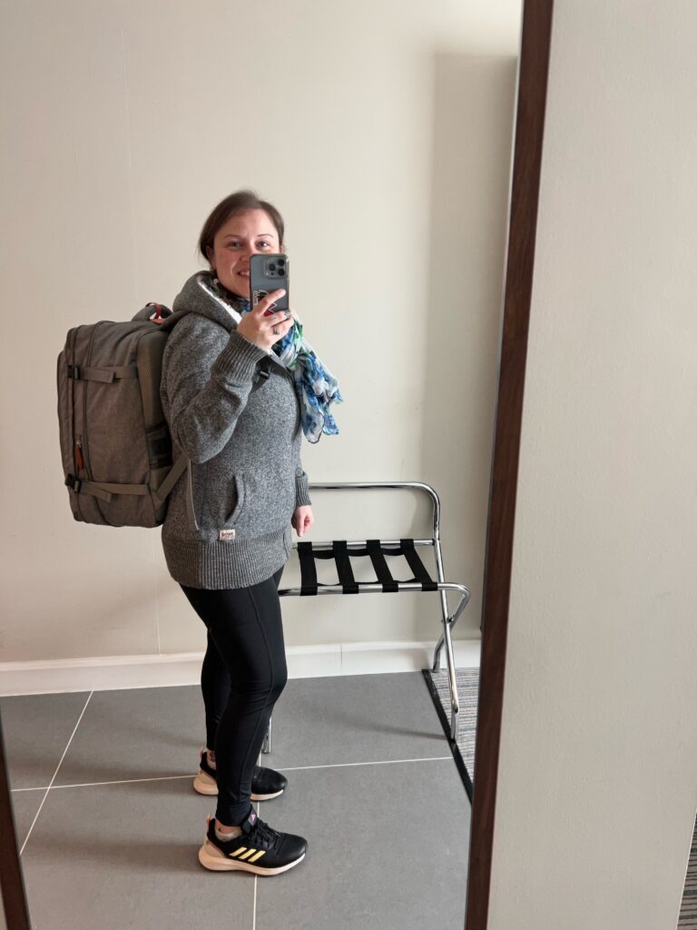 A lady is posing for a full length selfie, she has a large travel backpack on and is in a hotel room