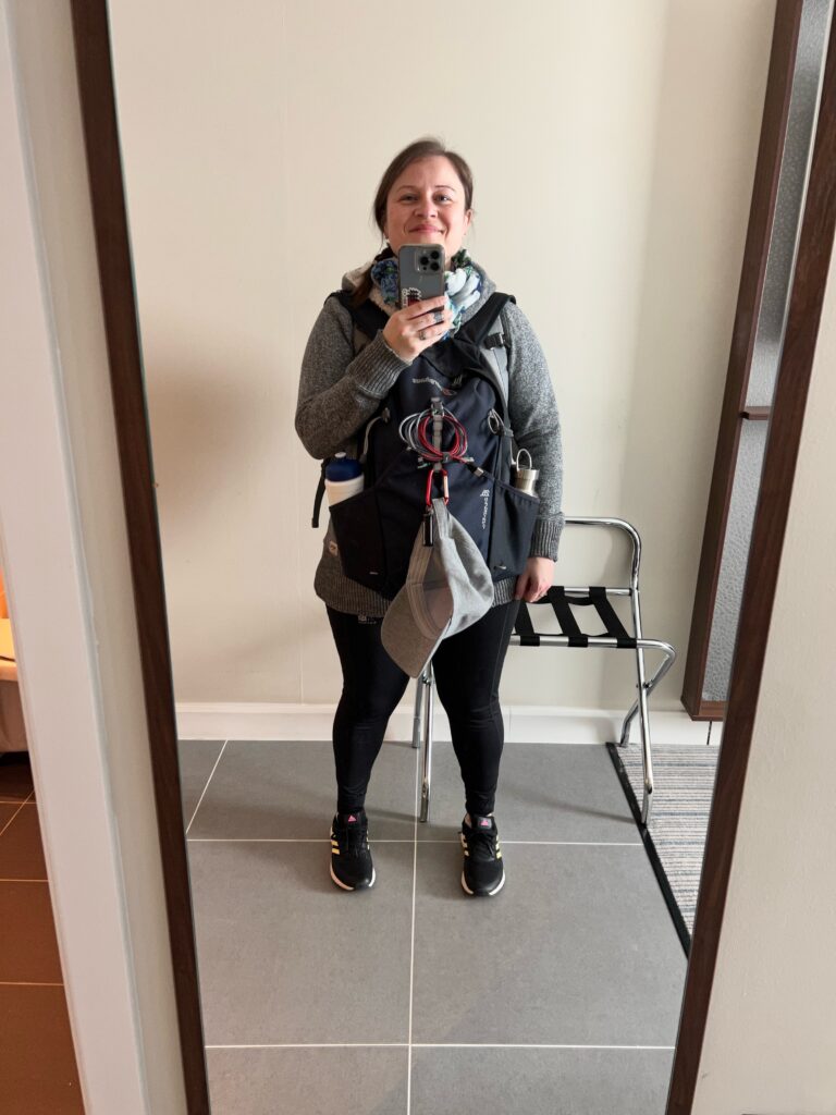 A lady is standing in a hotel room, taking a full length selfie in the mirror. She is visibly wearing two backpacks, a larger one on the back and a smaller one on her front with a baseball cap attached to it