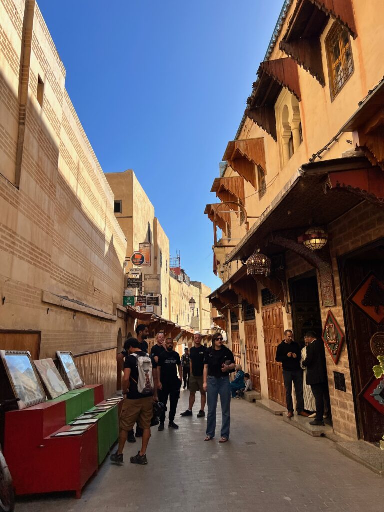 A group of people are stood in the narrow streets of Medina de Fez, local men are chatting in the doorway and there is a table of paintings opposite them